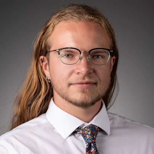 A picture of Keene Morrow, a non-binary adult with fair skin and long golden blonde hair wearing a white collared shirt, a multi-colored floral tie, and glasses. Keene hates this photo.